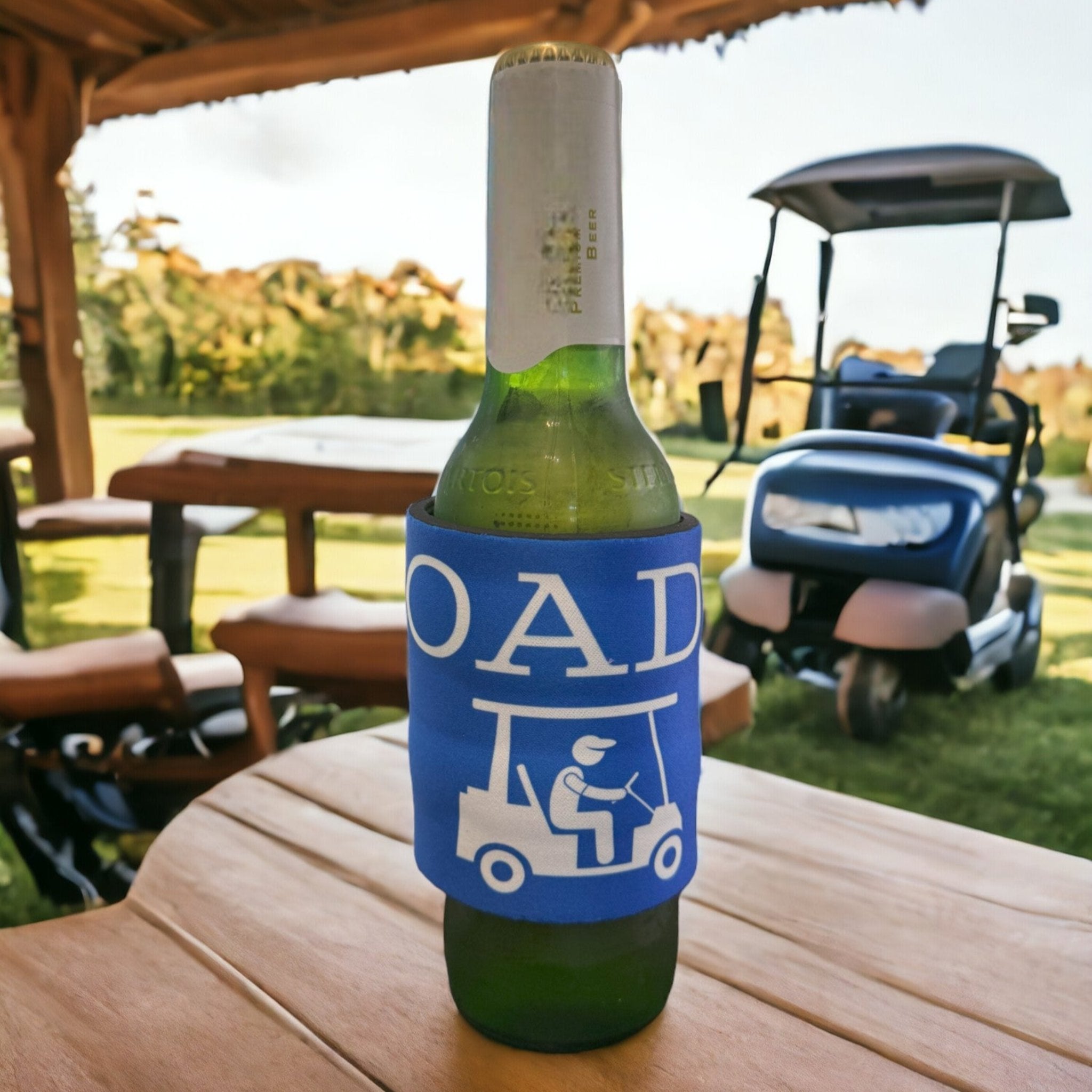 Blue custom drinkware sleeve with 'ROADIE' and golf cart design on a beer bottle, set on a wooden table at a golf course, showcasing unique promo items.