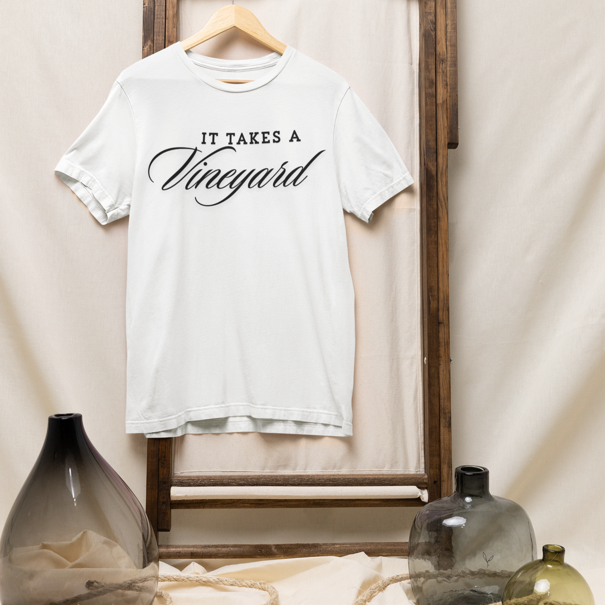 A "It Takes a Vineyard" Short Sleeve T-Shirt hangs on a wooden dowel. The dowel is attached to a rustic wooden frame standing against a beige fabric background, embodying the essence of comfort. Below the t-shirt are several glass vases in various colors and shapes, positioned on the floor, partially wrapped with a beige rope.