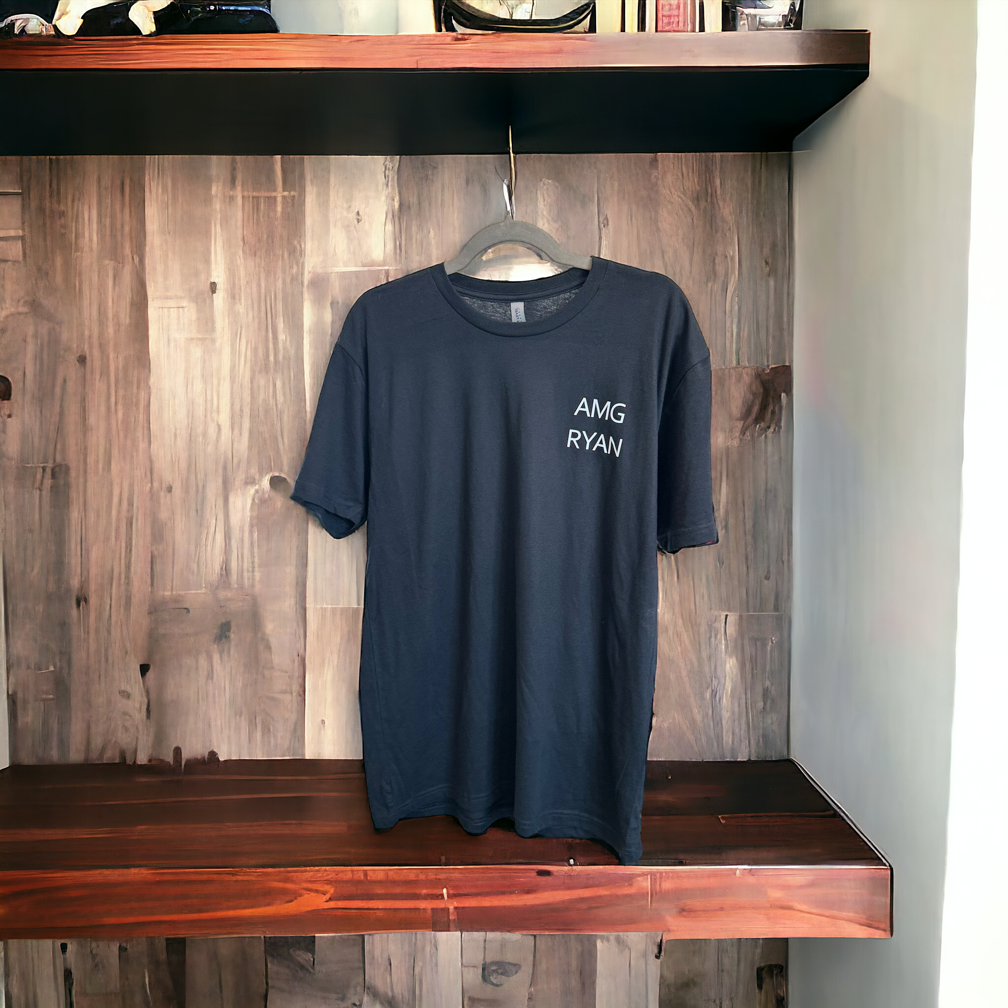 Black custom t-shirt with 'AMG RYAN' text, displayed on a wooden shelf against a rustic wood panel background, showcasing branded products