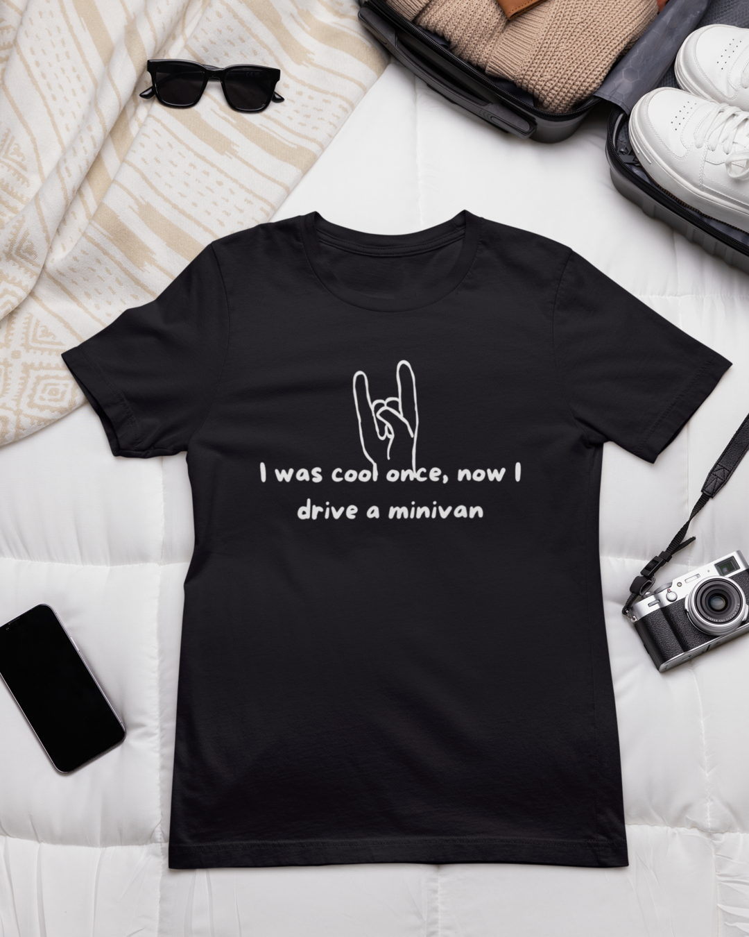 A "I Was Cool Once, Now I Drive a Minivan" Short Sleeve T-Shirt lies on a white quilted surface, accompanied by folded beige clothing, a pair of white sneakers, black sunglasses, a black smartphone, and an old-fashioned black camera. The shirt features a hand making a rock and roll sign and the text "I was cool once, now I drive a minivan.