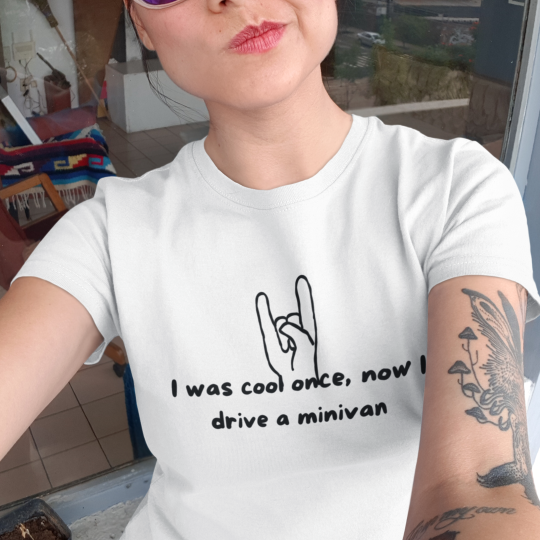 A woman is taking a selfie, wearing an "I Was Cool Once, Now I Drive a Minivan" Short Sleeve T-Shirt that has a hand showing the rock and roll sign with the text "I was cool once, now I drive a minivan." She has pink sunglasses and a tattoo of a butterfly on her right arm. The background shows a balcony with a chair and colorful blanket.