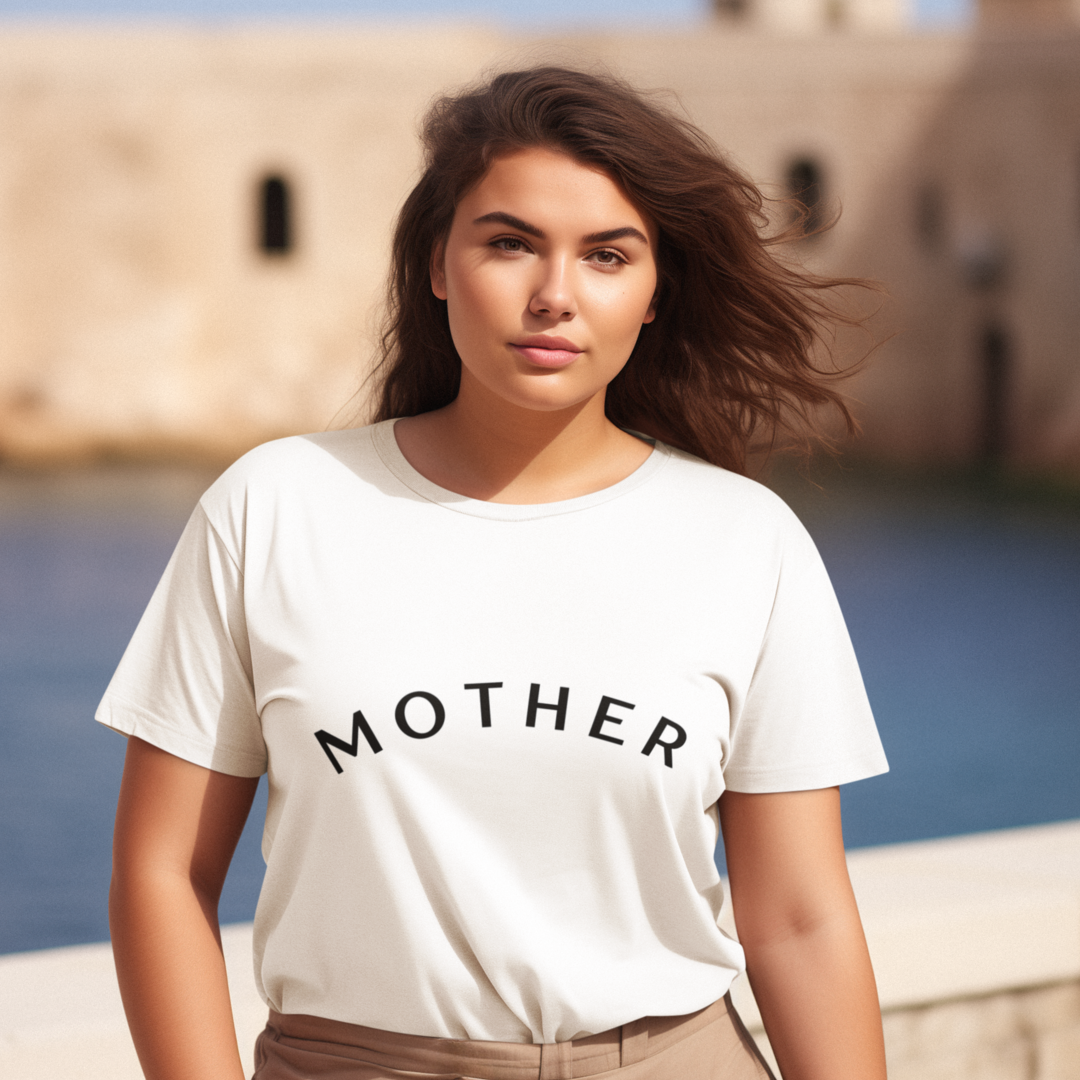 A woman with long brown hair stands outdoors in front of a body of water and stone buildings. She wears a chic and minimalist white "Mother" Short Sleeve T-Shirt with the word “MOTHER” printed in bold black letters. The wind blows her hair gently, and she gazes directly at the camera with a neutral expression.