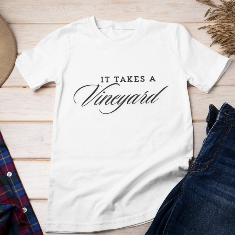 A "It Takes a Vineyard" Short Sleeve T-Shirt with the phrase "It Takes a Vineyard" in black script, styled in a calligraphic font, is laid out flat on a light wooden surface. Surrounding the shirt are a brown hat, a sprig of dried wheat, and comfort essentials: a red and blue plaid shirt and blue denim jeans.