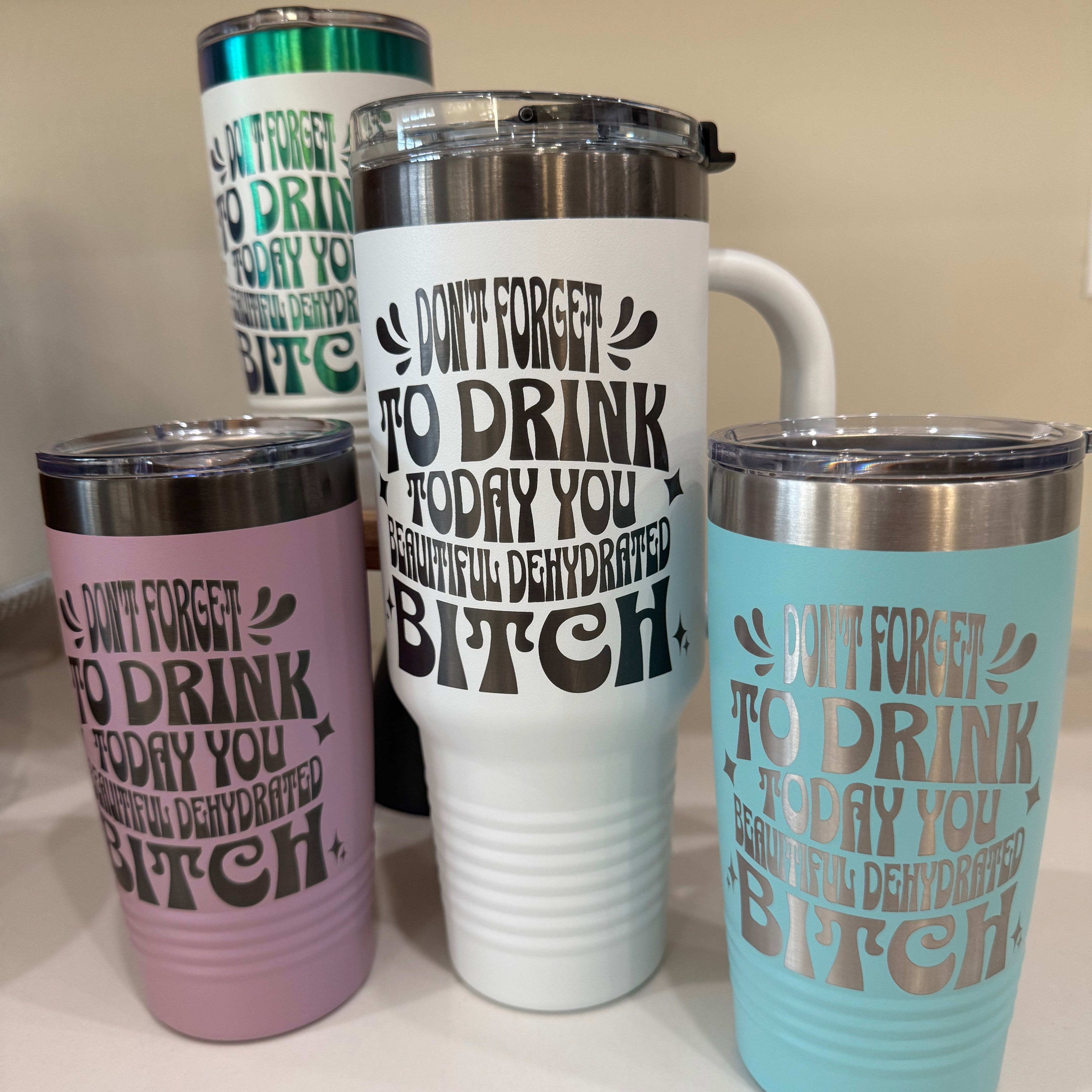 A collection of colorful insulated tumblers with humorous text that reads, "Don't forget to drink today, you beautiful dehydrated bitch." The tumblers come in white, lilac, turquoise, and green, designed for both hydration and style.