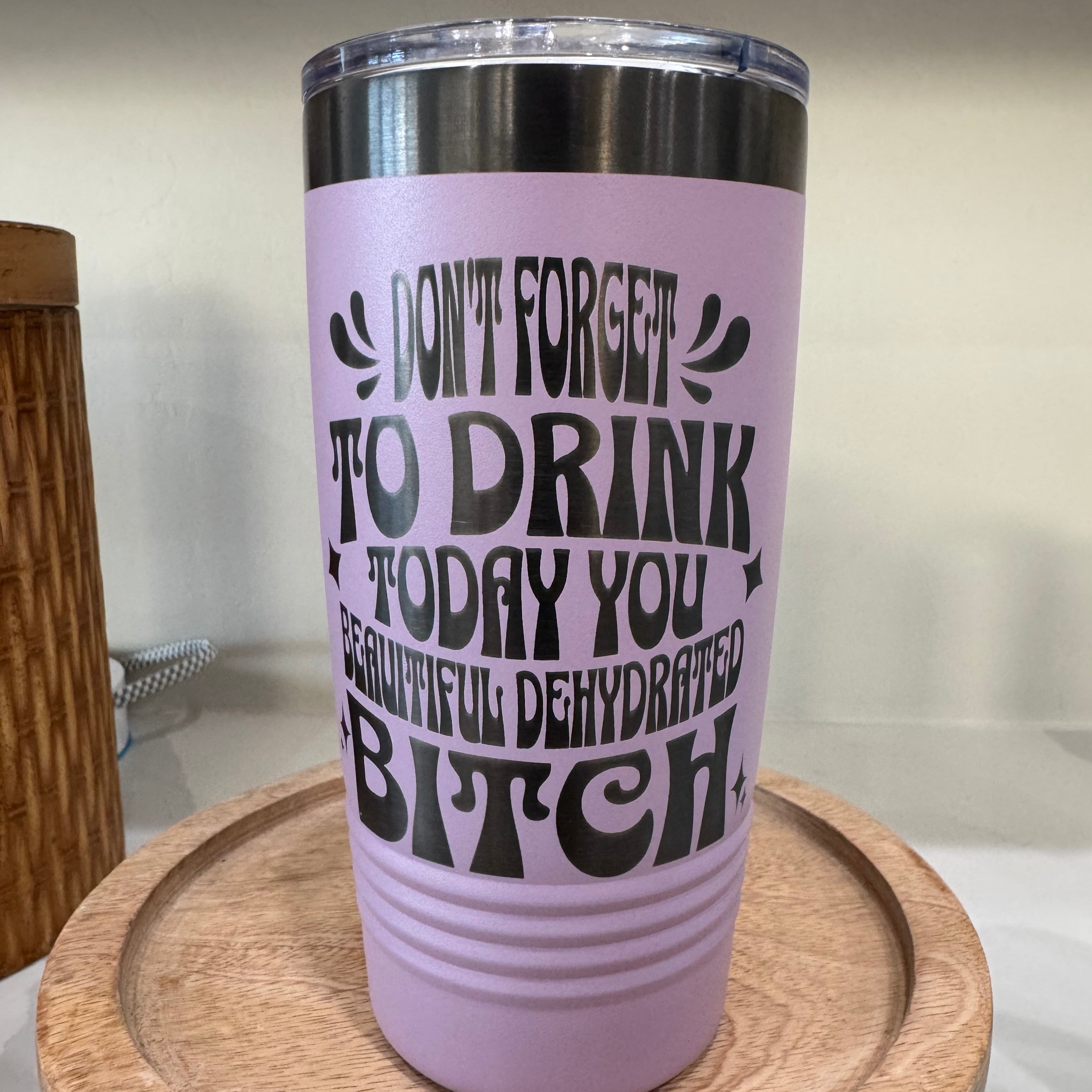 A lilac insulated tumbler with laser etched text that humorously reads, "Don't forget to drink today, you beautiful dehydrated bitch." The tumbler features a metallic green lid and sits on a wooden tray.