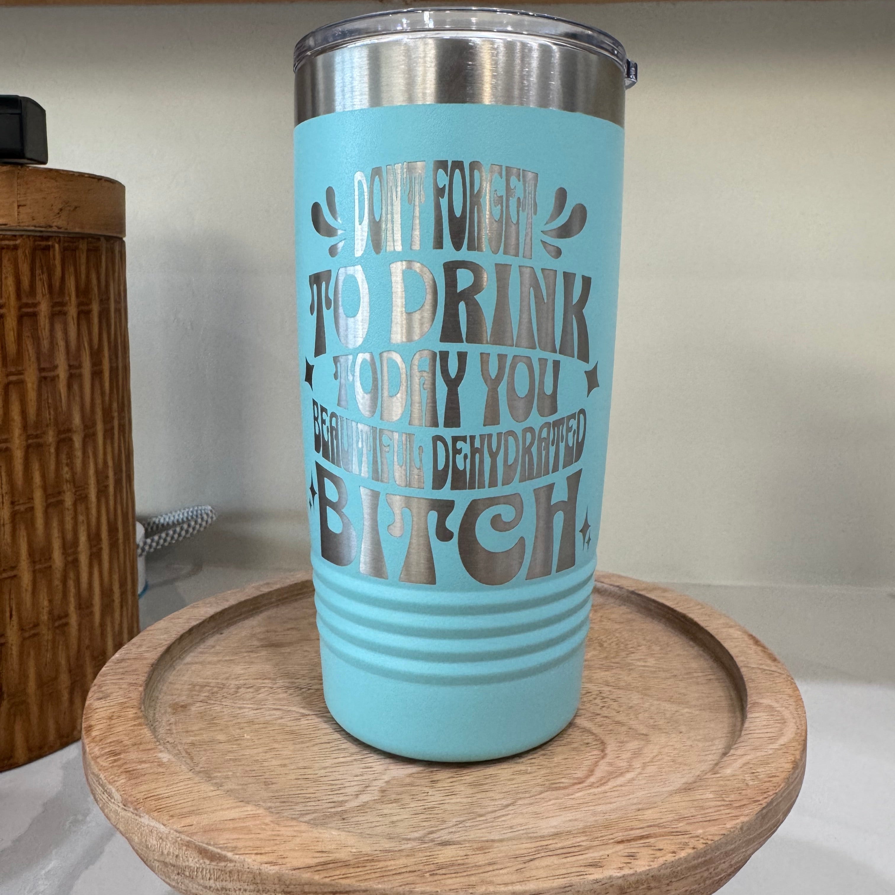 A teal insulated tumbler with laser etched text  that humorously reads, "Don't forget to drink today, you beautiful dehydrated bitch." The tumbler features a metallic green lid and sits on a wooden tray.