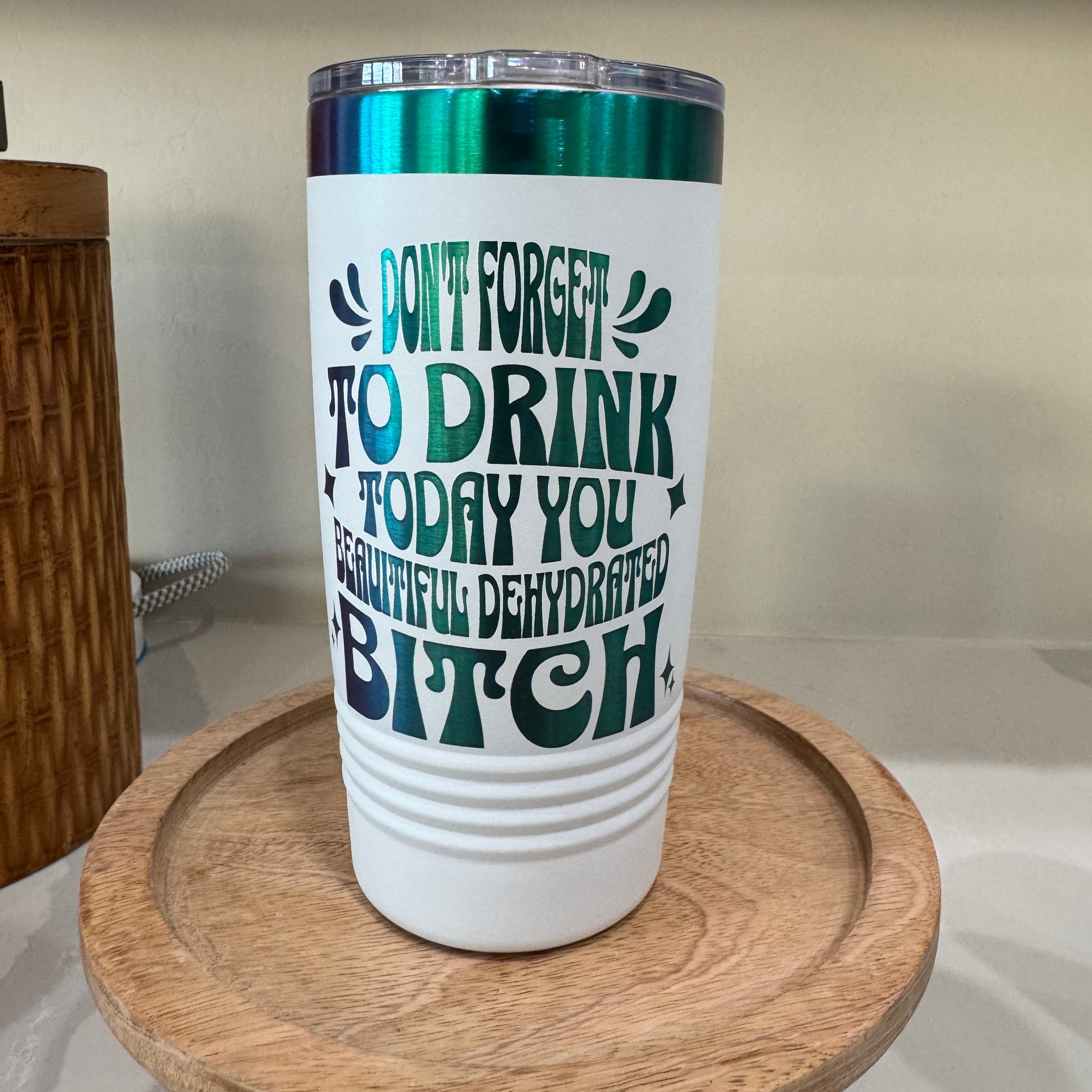 Image of a white insulated tumbler with green and black text that humorously reads, "Don't forget to drink today, you beautiful dehydrated bitch." The tumbler features a metallic green lid and sits on a wooden tray.