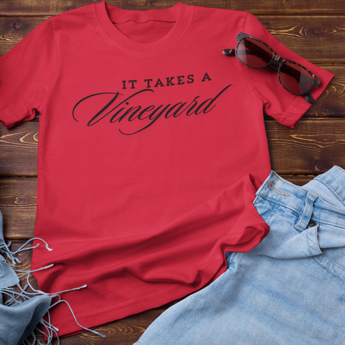 A "It Takes a Vineyard" Short Sleeve T-Shirt with the text "IT TAKES A Vineyard" written in black script is laid out on a dark wooden surface. Surrounding the T-shirt are a pair of sunglasses, a light blue scarf with fringes, and a pair of light blue denim shorts—perfect for effortless comfort and style while celebrating motherhood.
