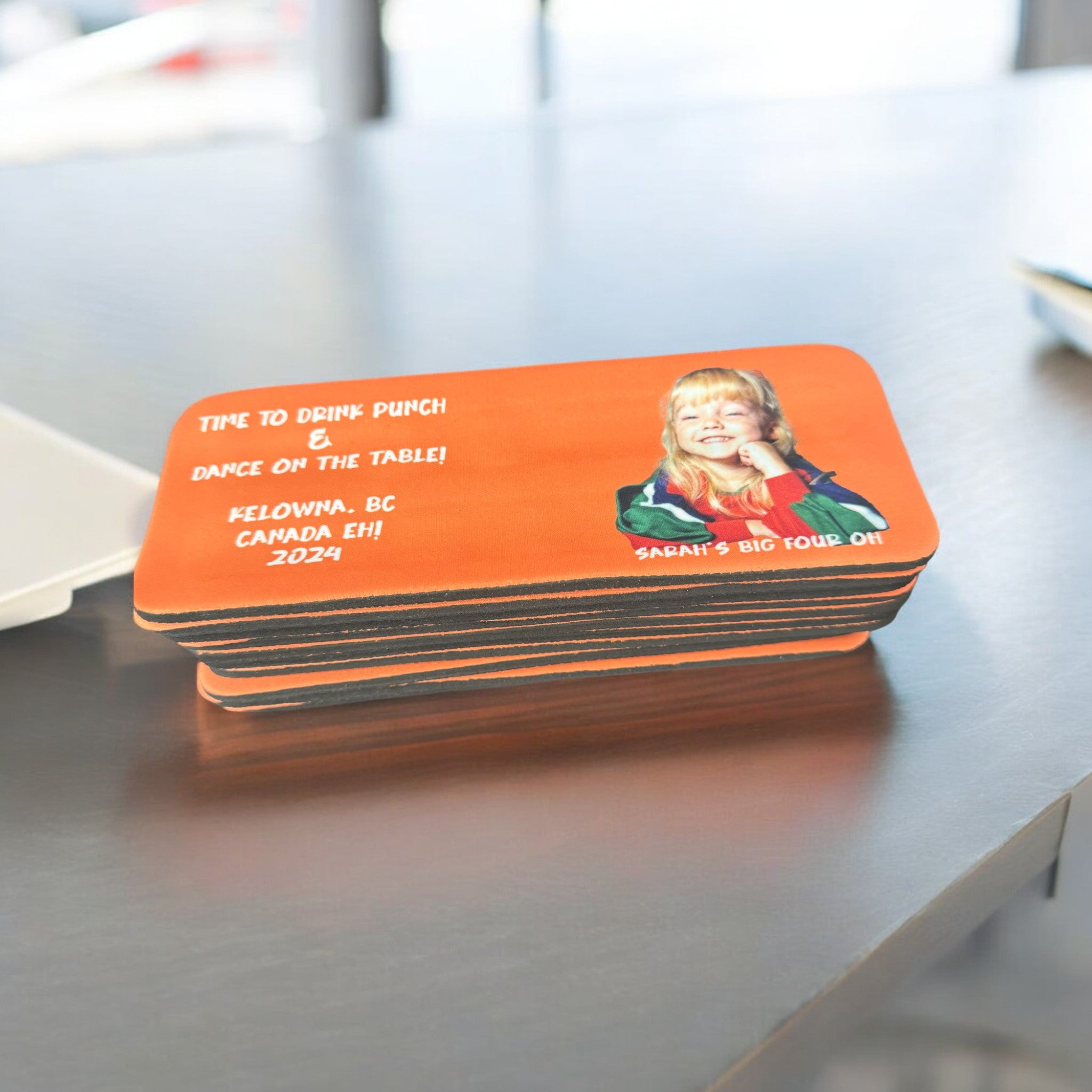 Stack of orange slap wrap drink sleeves featuring a childhood photo of a smiling blonde girl in a colorful jacket. The text reads "Time to drink punch & dance on the table! Kelowna, BC Canada Eh! 2024" with "Sarah's Big Four Oh!" written at the bottom right. The sleeves are stacked on a reflective surface indoors.