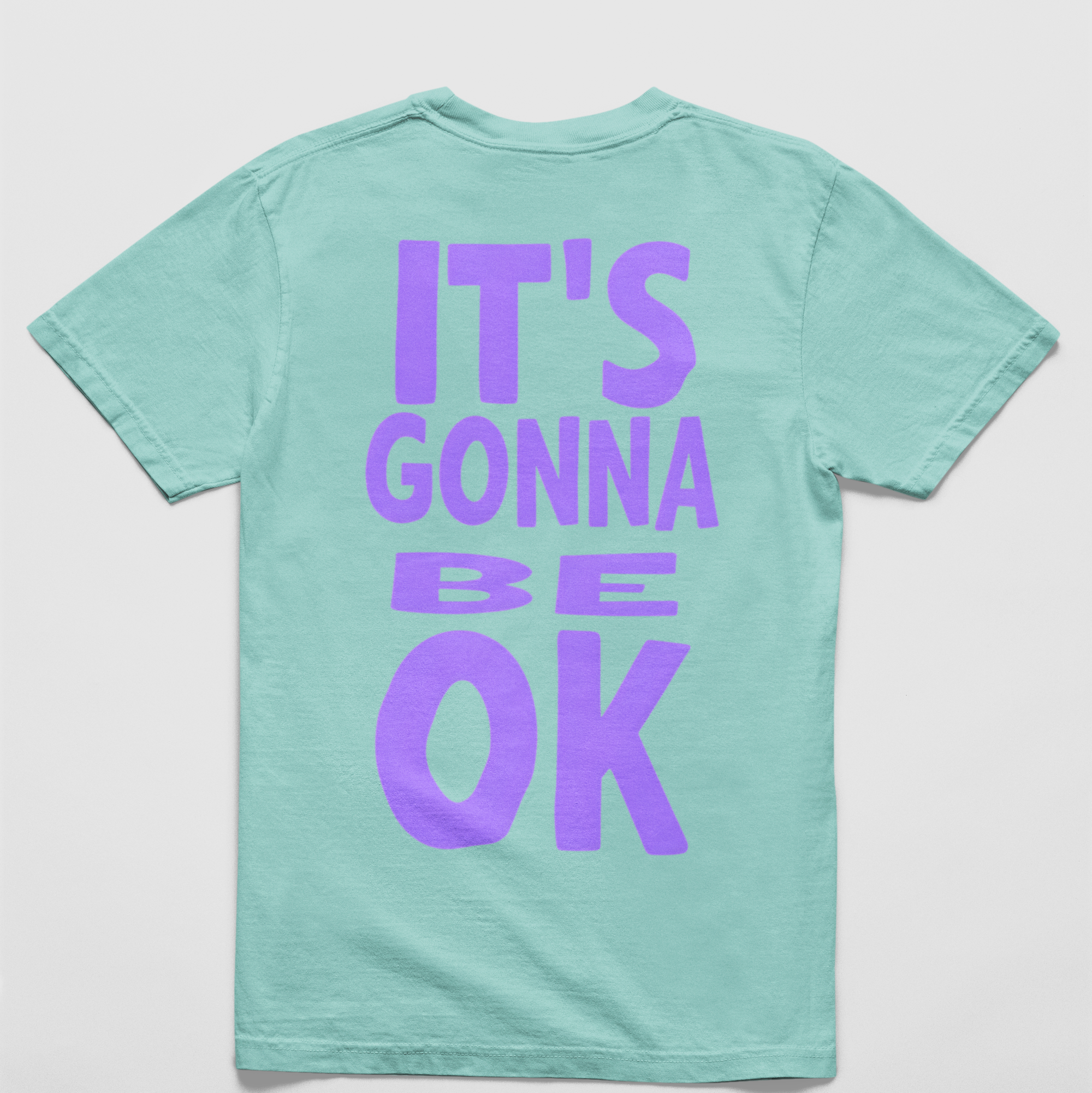 Oversized chalky mint tee with "It’s Gonna Be OK" printed in bold purple text on the back, styled for casual and confident comfort.