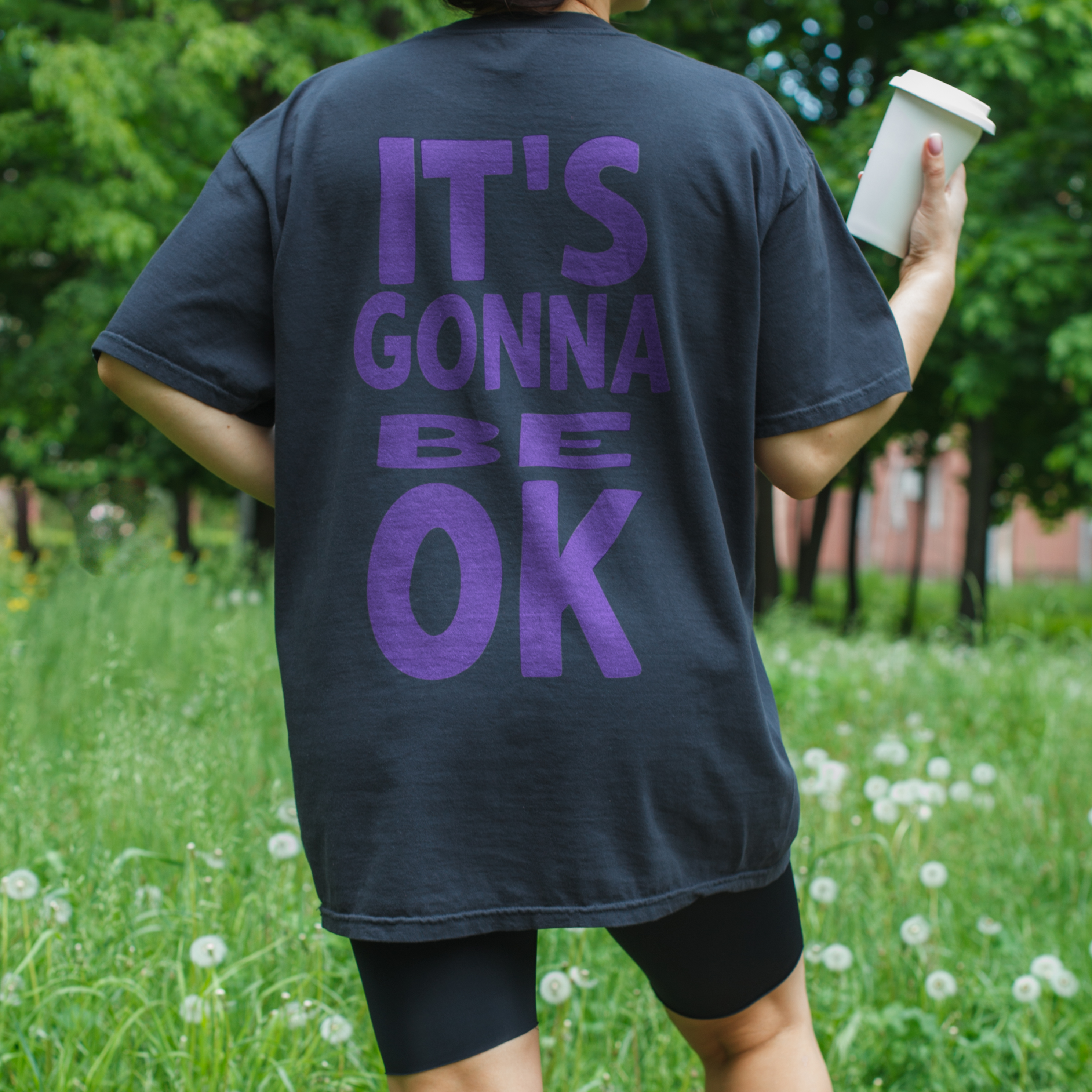 woman wearing Oversized black tee with "It’s Gonna Be OK" printed in bold purple text on the back, styled for casual and confident comfort.