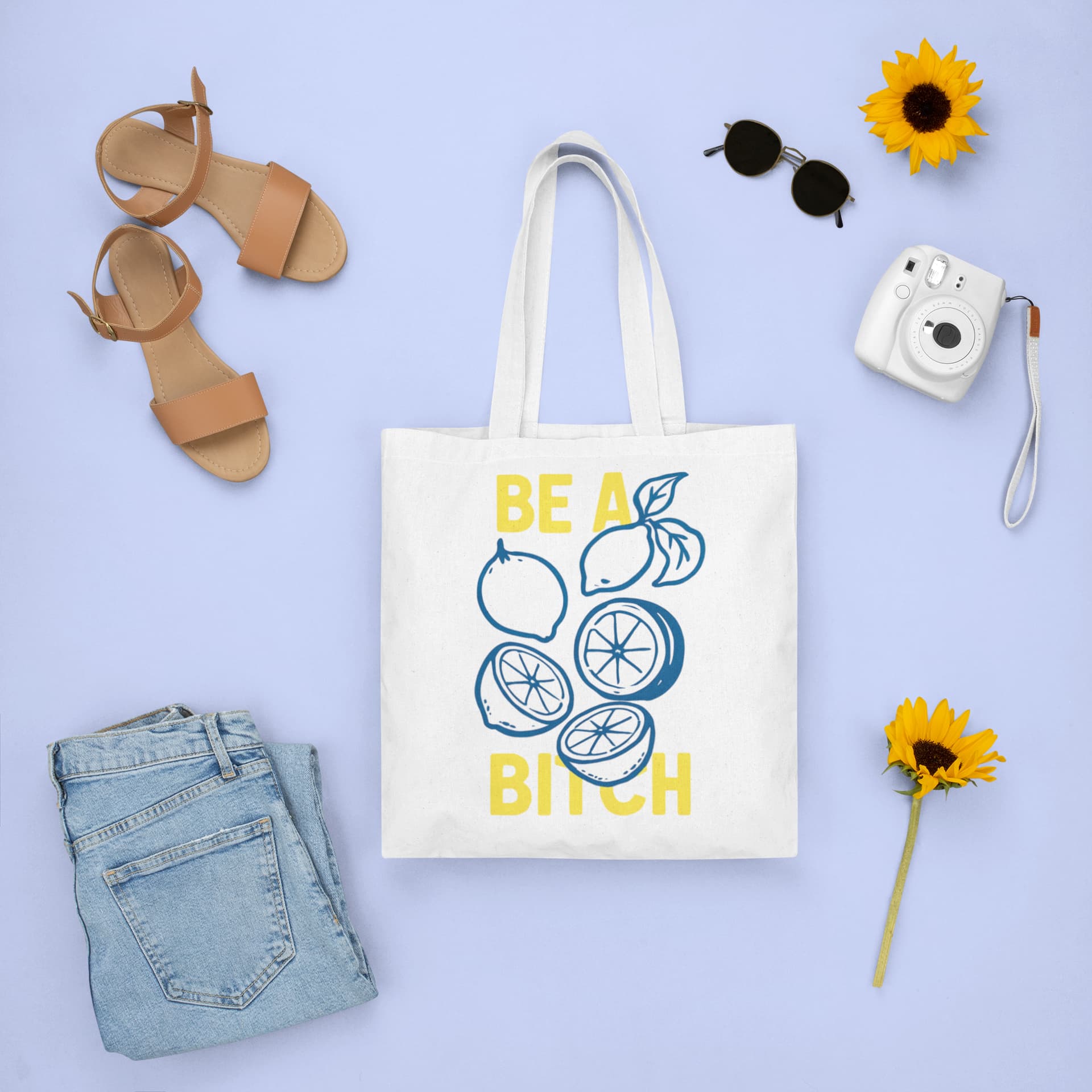 Tote bag with lemon design and text, surrounded by sandals, jeans, camera, and sunflowers on a light blue background