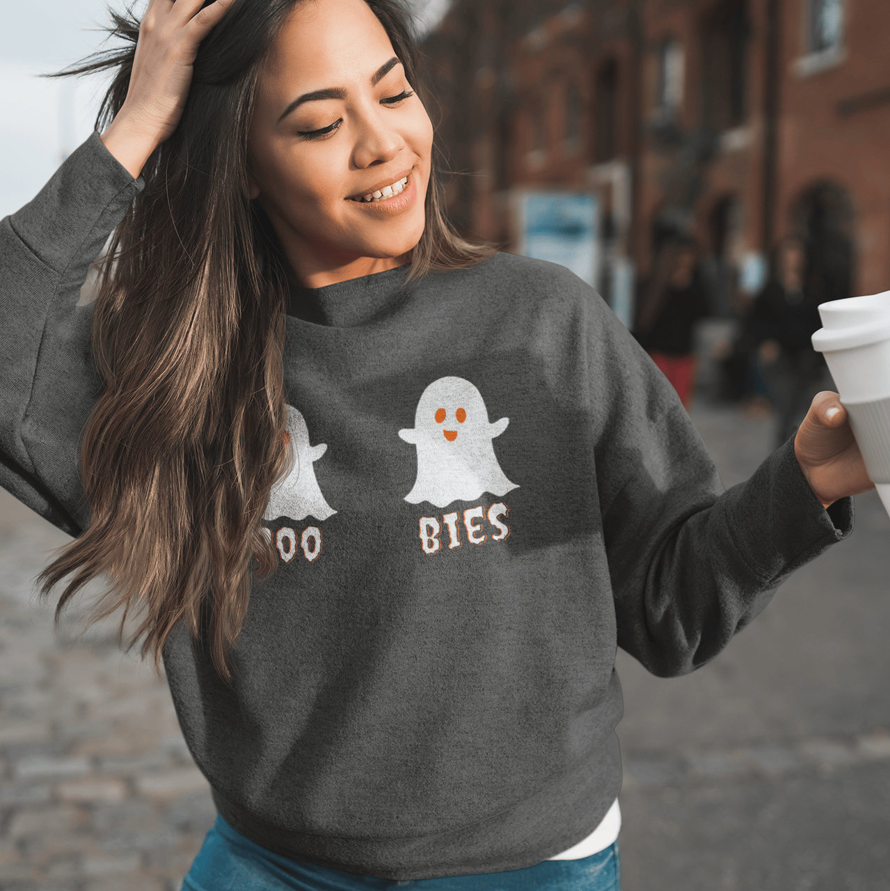 Woman wearing a dark gray cozy Halloween sweatshirt with two cute ghost illustrations and the playful text "BOO BIES," holding a coffee cup outdoors, perfect for casual festive outings.