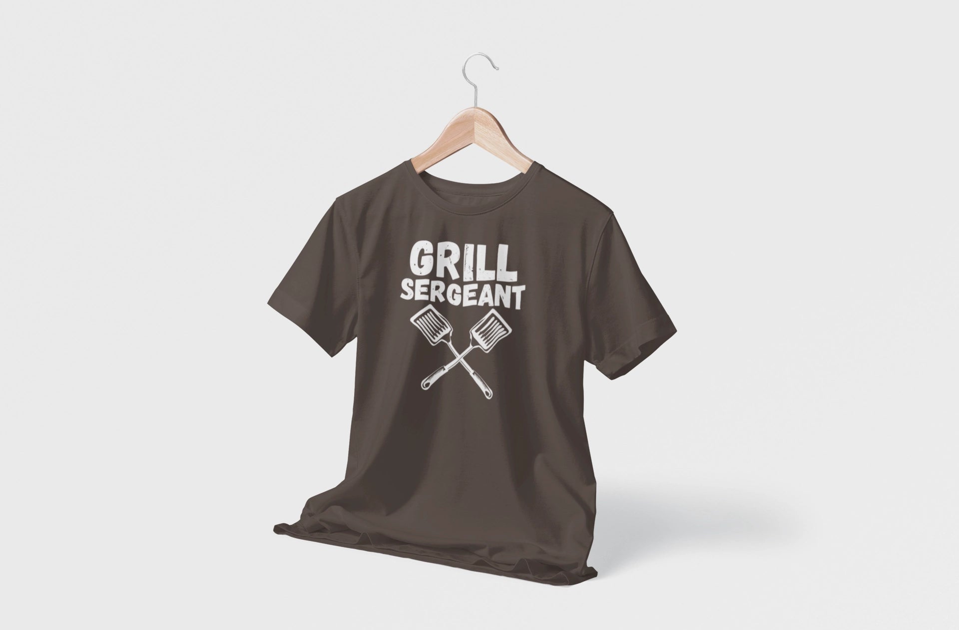 Brown t-shirt with 'GRILL SERGEANT' text and crossed grill tools on a hanger against a white background