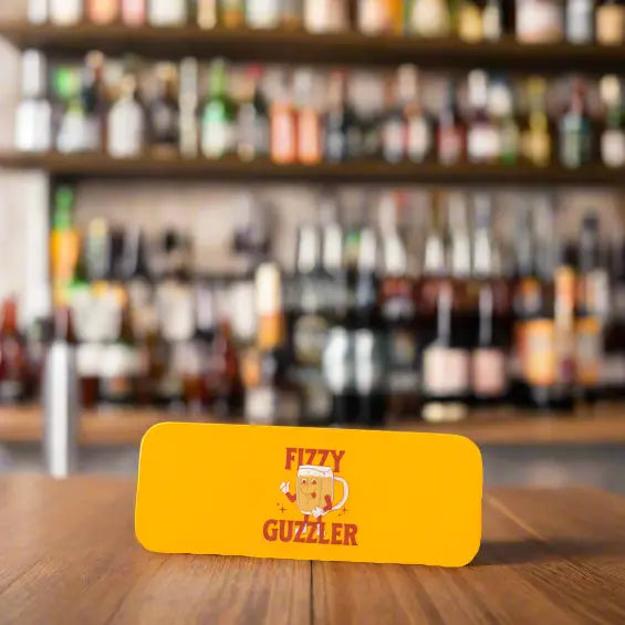 Fizzy Guzzler slap wrap koozie with beer mug graphic shown on a bar table with blurred bottles in the background