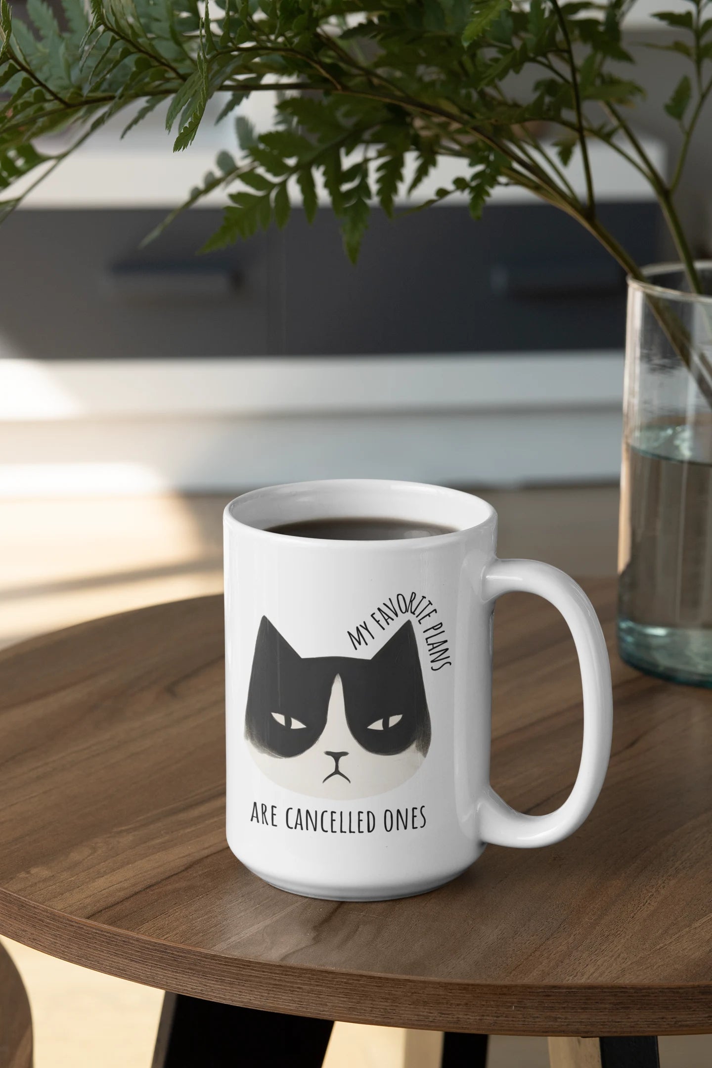 White mug with a black cat illustration and text on a wooden table with a plant in the background