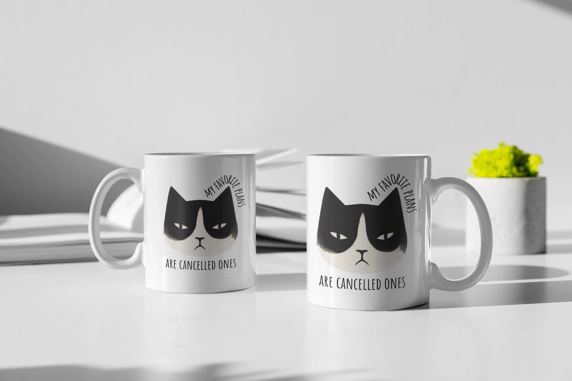 Two white mugs with black cat illustrations and text on a light surface.