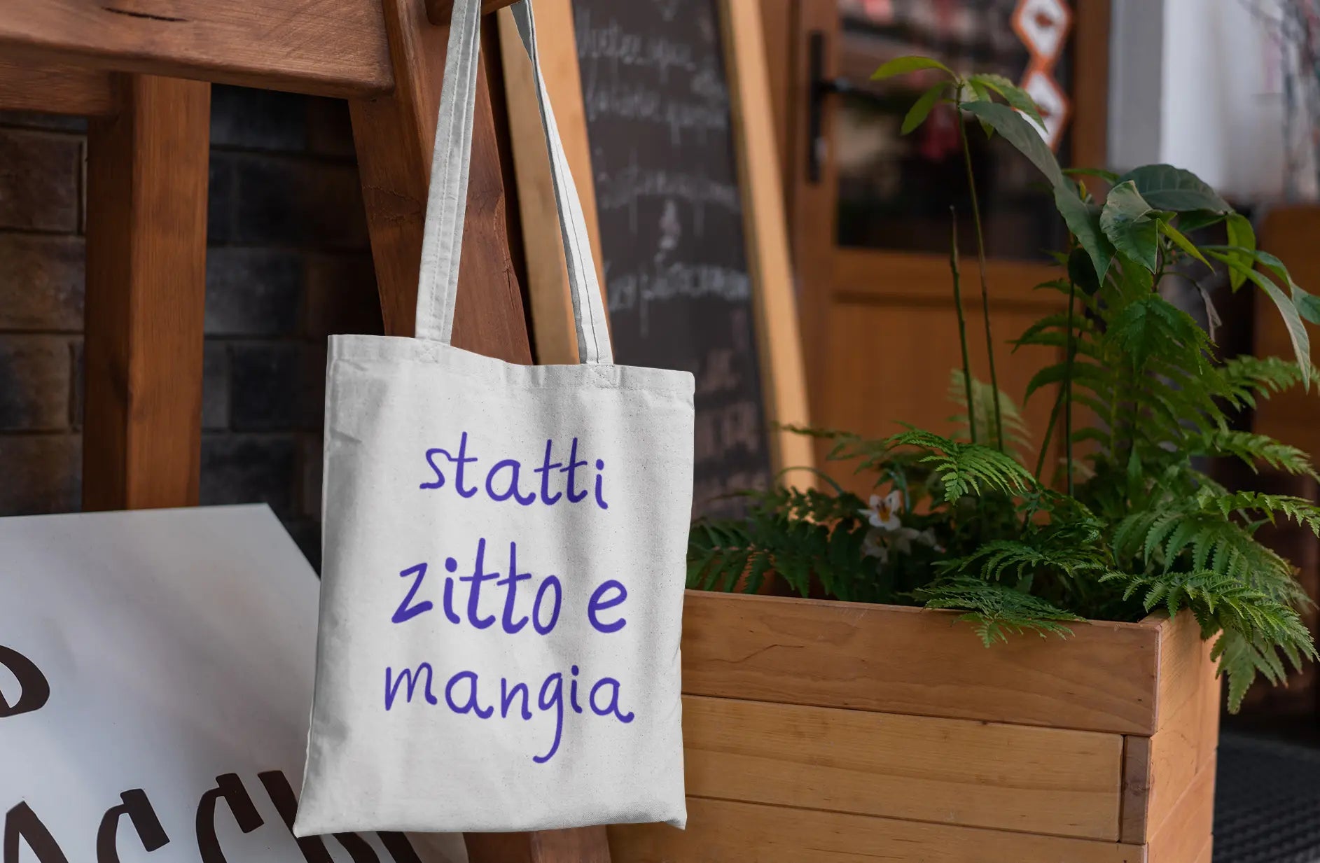 White tote bag with Italian text 'statti zitto e mangia' hanging on a wooden post outdoors.