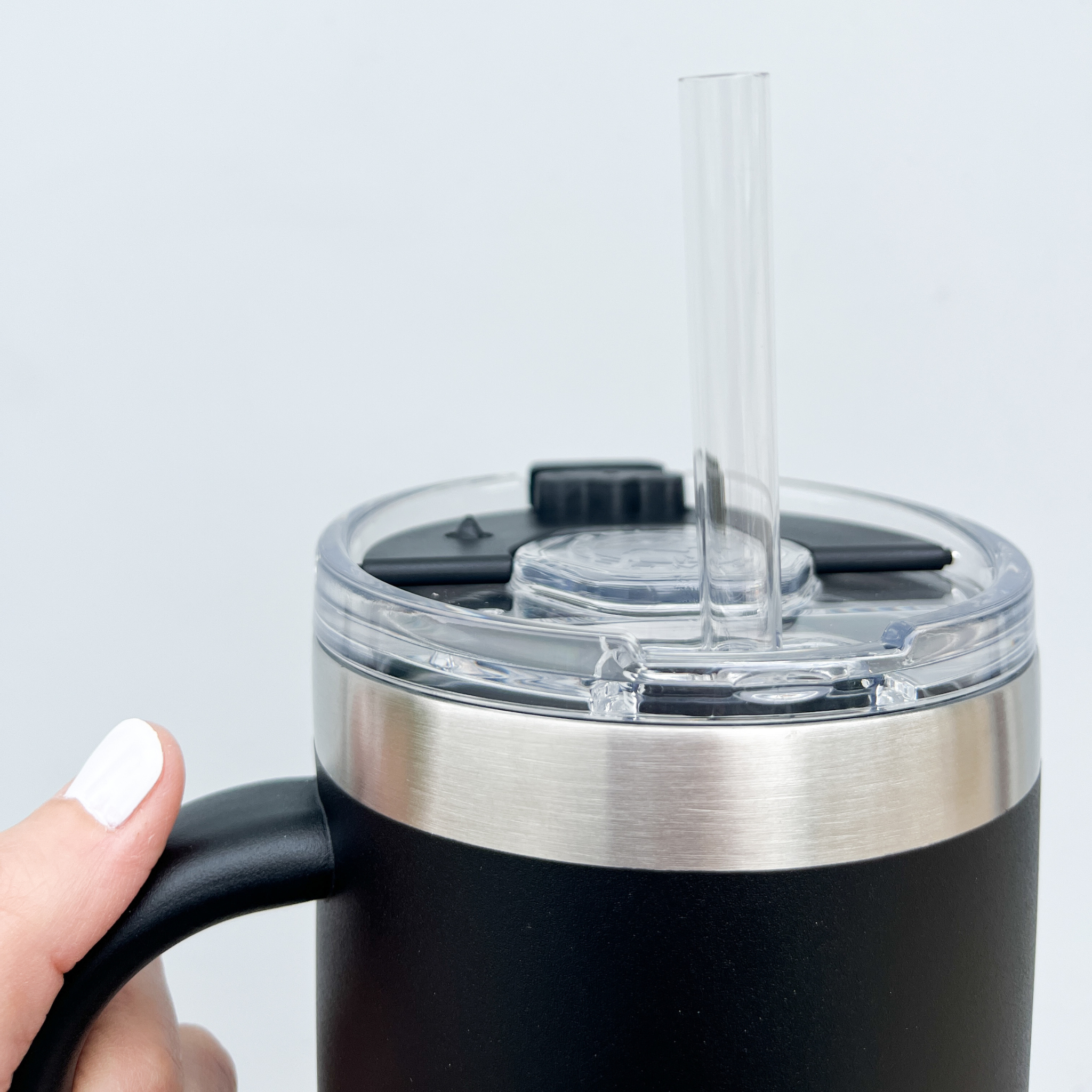 Close-up of a black tumbler with a clear lid, featuring a built-in straw and stainless steel rim, held by a hand with white-painted nails.