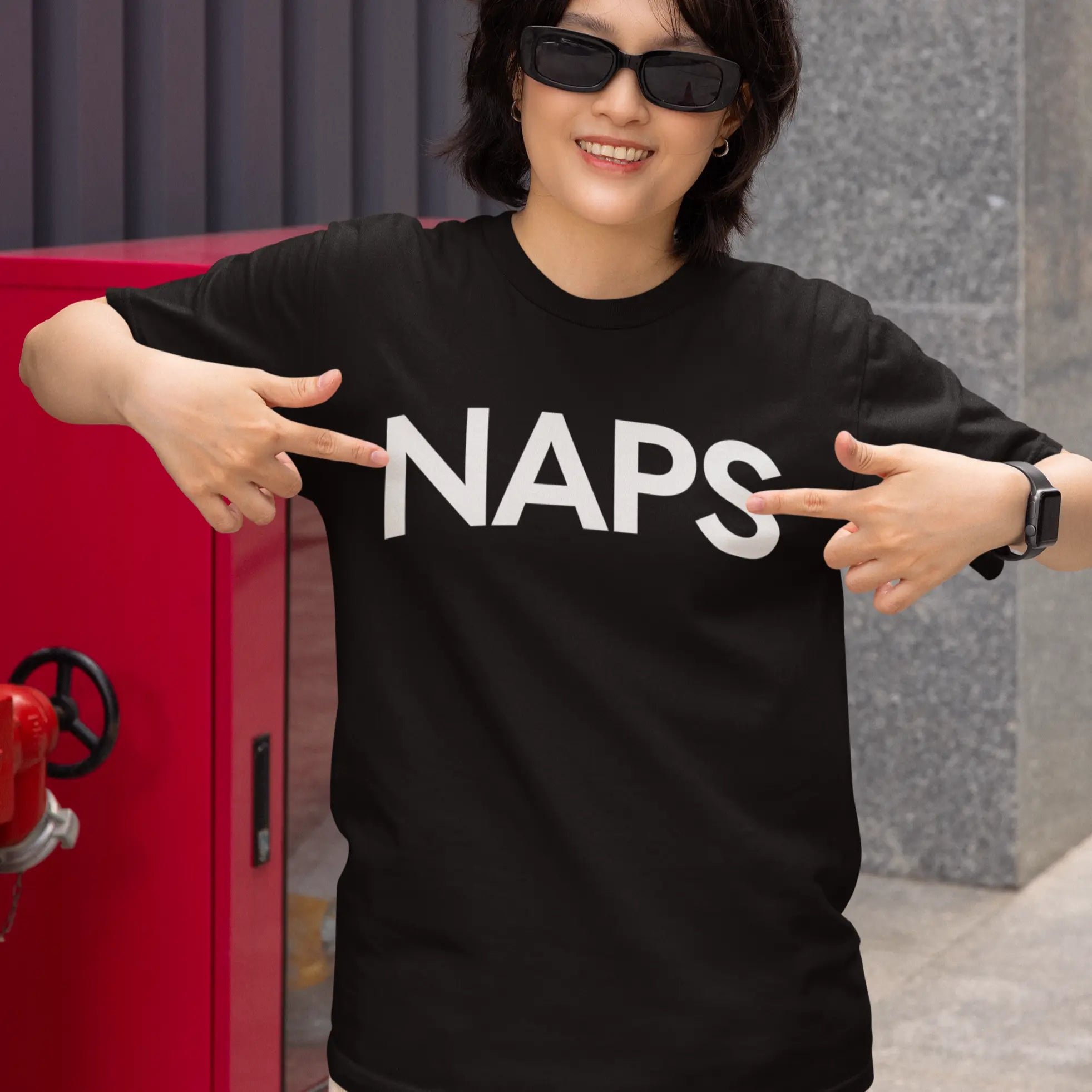 Person wearing a black t-shirt with 'NAPS' text, pointing at it, with a red object and gray wall in the background.