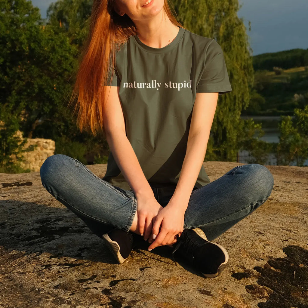 Woman sitting outdoors wearing “naturally stupid” graphic t-shirt in a nature lifestyle mockup