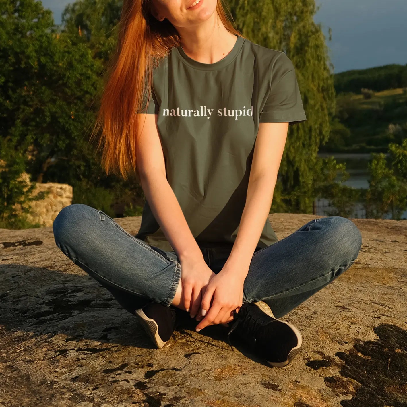 Woman sitting outdoors wearing “naturally stupid” graphic t-shirt in a nature lifestyle mockup
