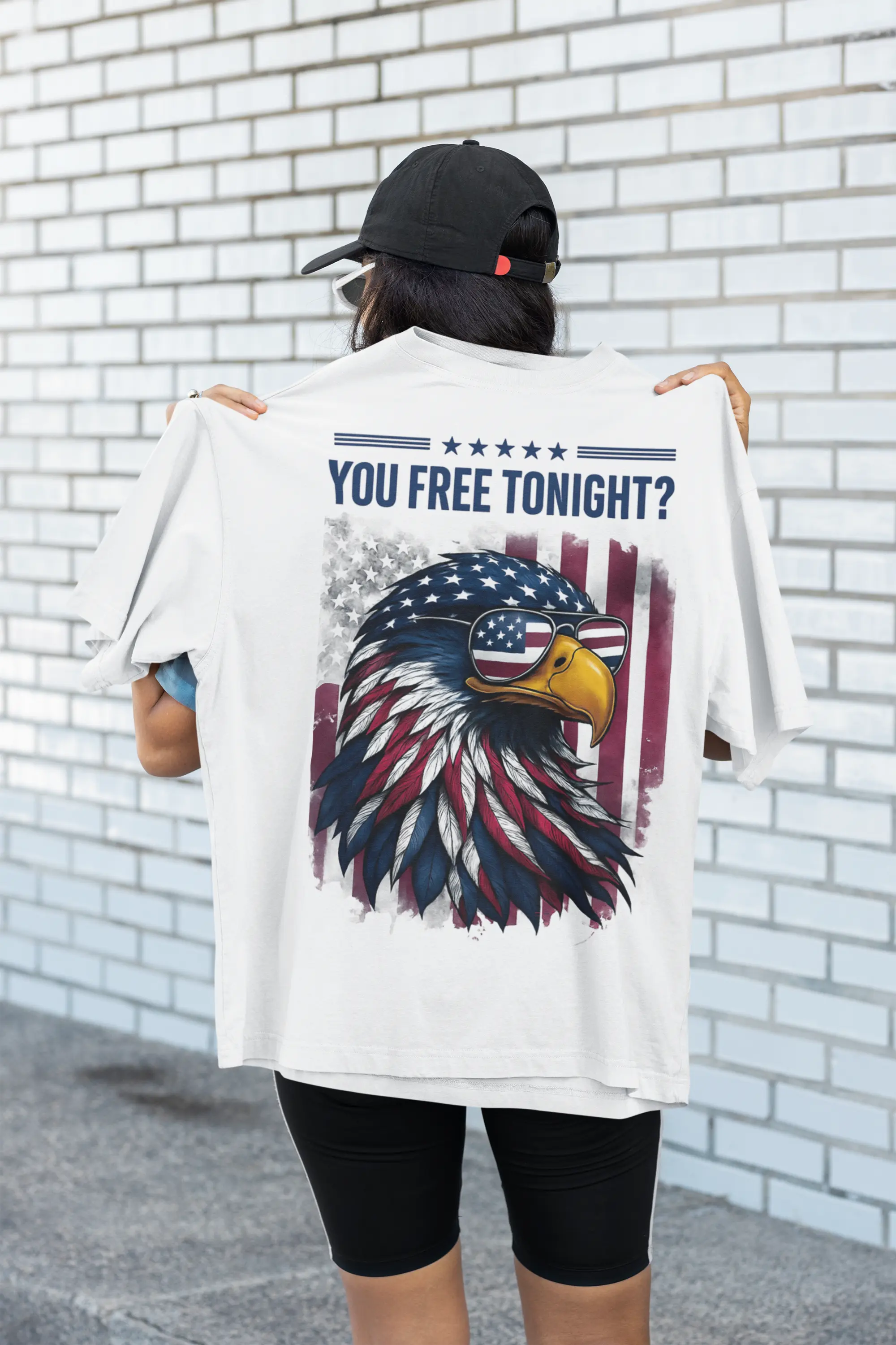 Person wearing a white t-shirt with an eagle graphic and text, standing against a white brick wall.