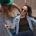 Two girls playing with shopping cart, one wearing black "Crown Straightener" t-shirt with red text - fun lifestyle moment celebrating friendship and good times