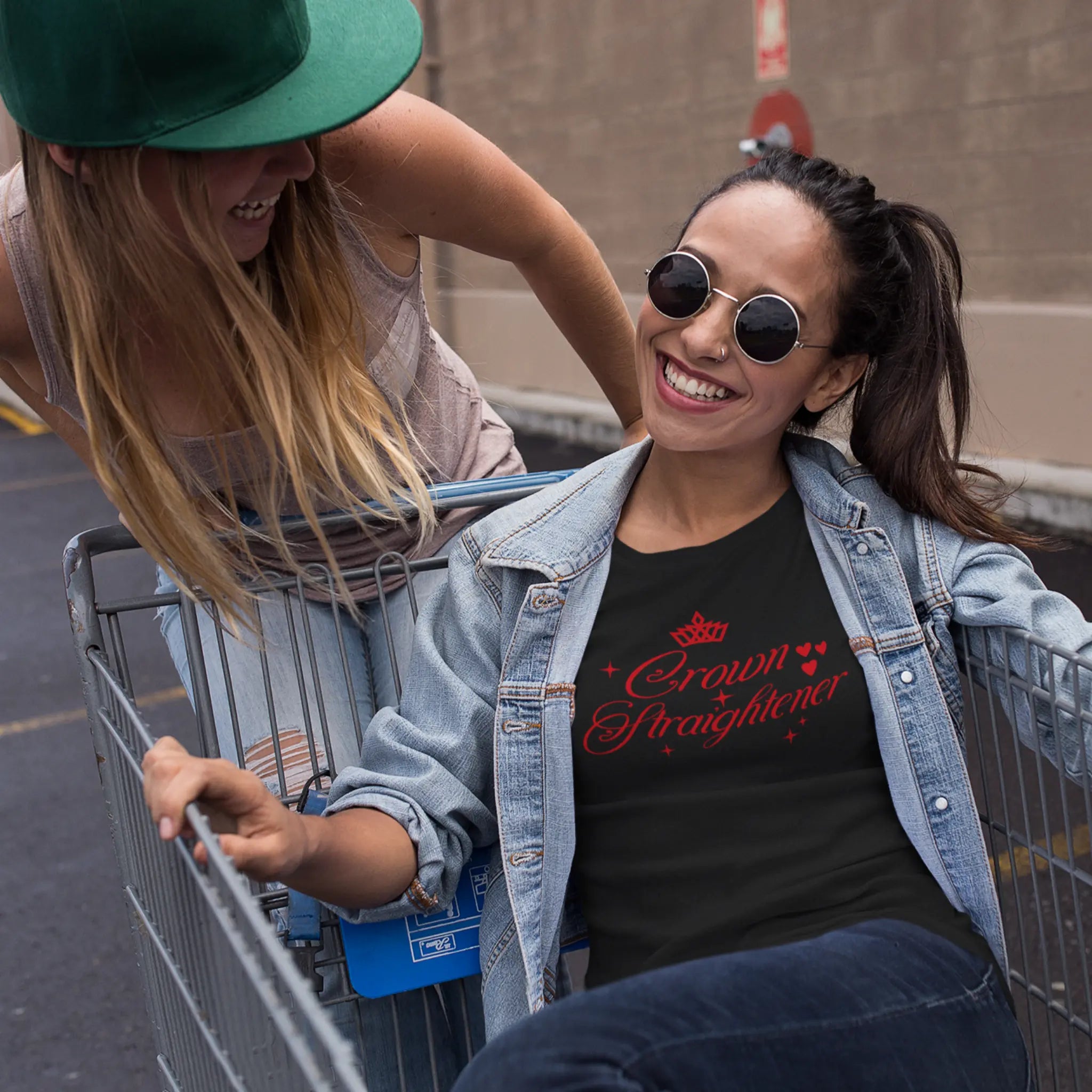 Two girls playing with shopping cart, one wearing black "Crown Straightener" t-shirt with red text - fun lifestyle moment celebrating friendship and good times