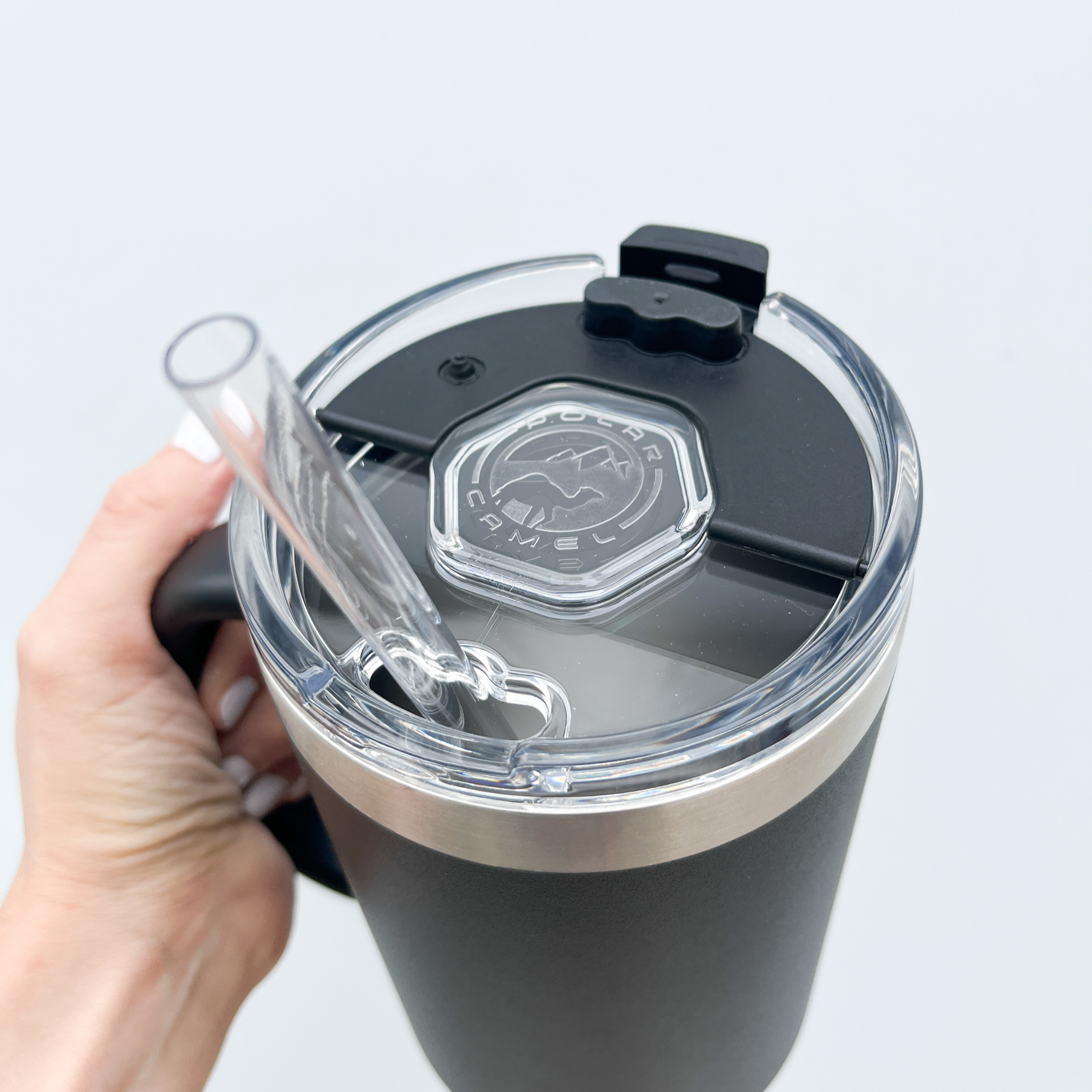 Close-up angle of a black tumbler with a clear lid, featuring a built-in straw and stainless steel rim, held by a hand with white-painted nails.