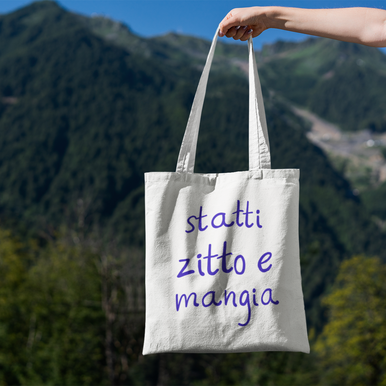 Canvas tote bag with purple text reading “statti zitto e mangia” on woman’s shoulder, hand holding straps

