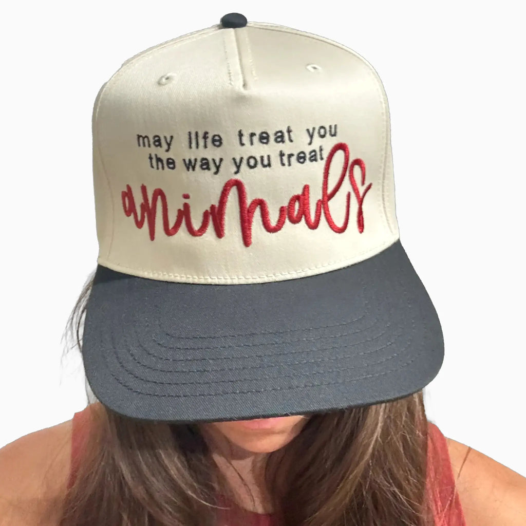 A person wearing a two-tone snapback cap with an off-white crown and a dark gray brim. The front of the cap features the embroidered text 'may life treat you the way you treat animals' in black and red script.