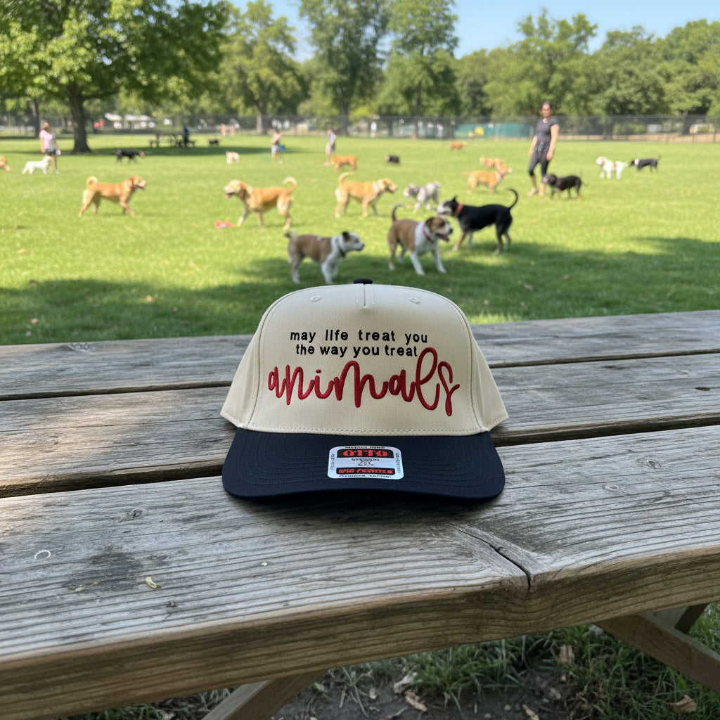 A two-toned snapback cap with embroidered text reading 'may life treat you the way you treat animals' in black and red script on the front