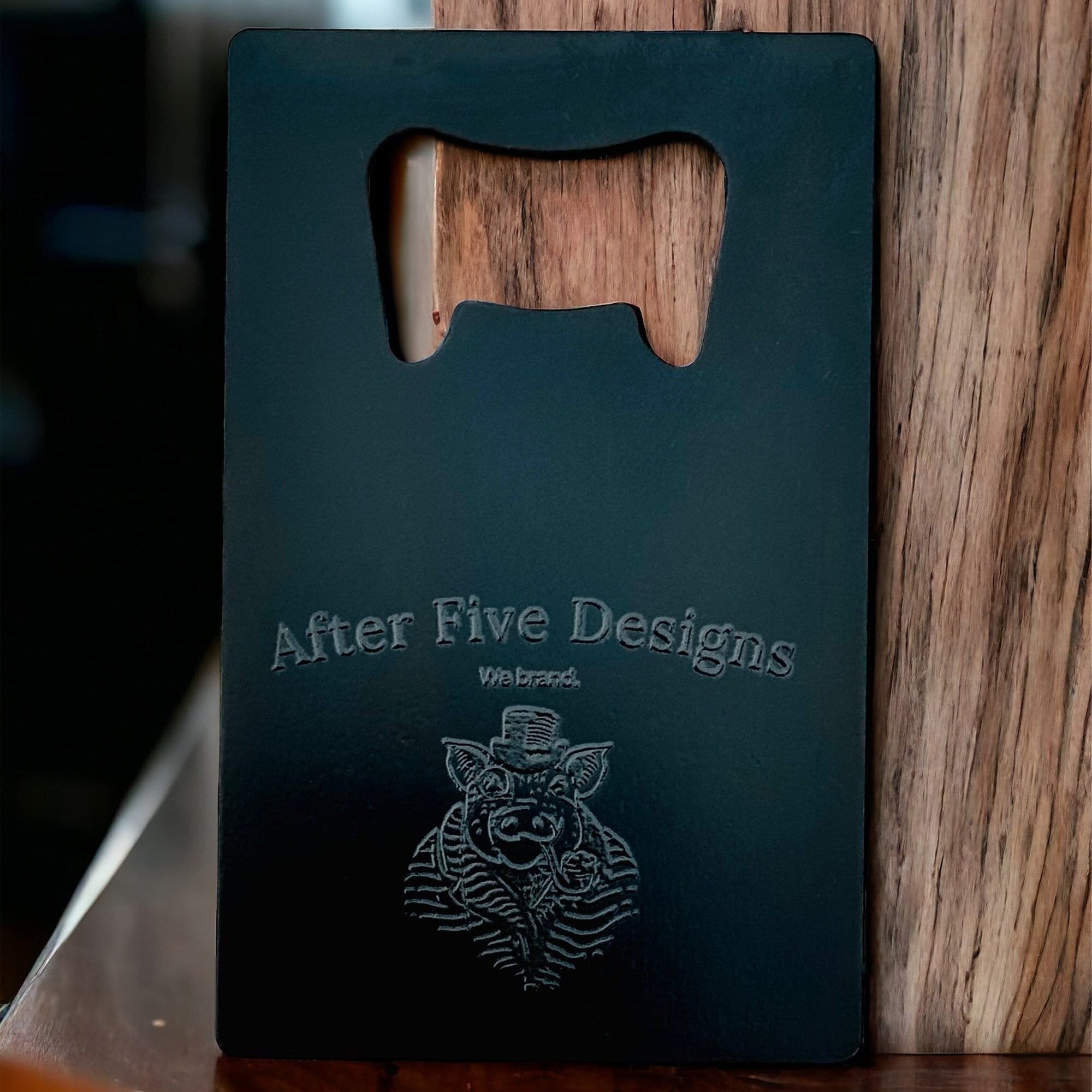 Black custom bottle opener business card engraved with "After Five Designs" logo, featuring a pig in a top hat, designed for branding purposes.