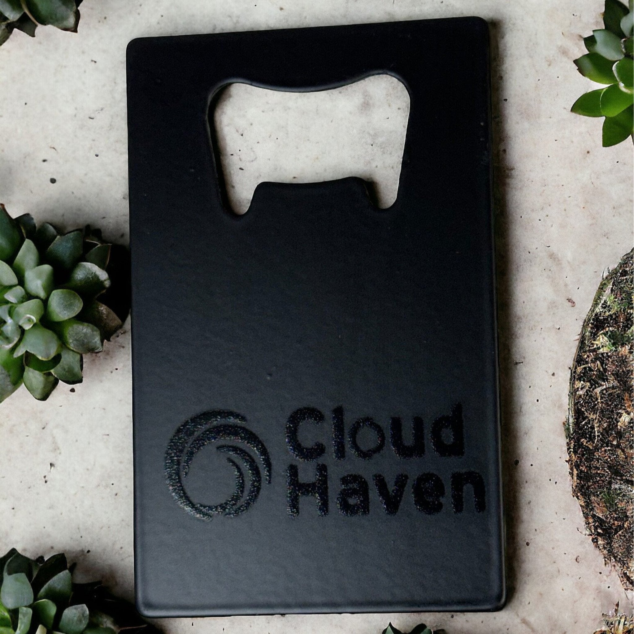 Black custom bottle opener business card with "Cloud Haven" logo, designed for eco-friendly branding and promotional products, surrounded by small succulents on a concrete surface.